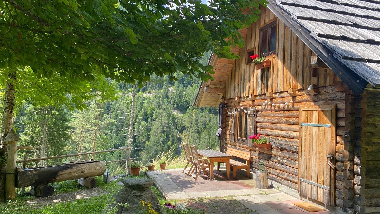 Die Außenseite des Bed and Breakfast mit einer Holzfassade, einem gepflasterten Sitzbereich im Freien und Blick auf die bewaldete Berglandschaft.