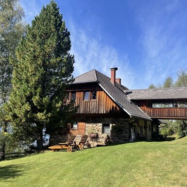 Das Äußere der Alm mit Holz- und Steinfassade, einem Verbindungssteg, der umliegenden Grünfläche und Holzliegestühlen.
