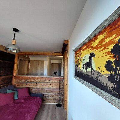 Der Innenbereich der Alm mit einem Schlafsofa, Holzwänden, einer Hängelampe und einem gerahmten Wandteppich mit Pferdemotiv.