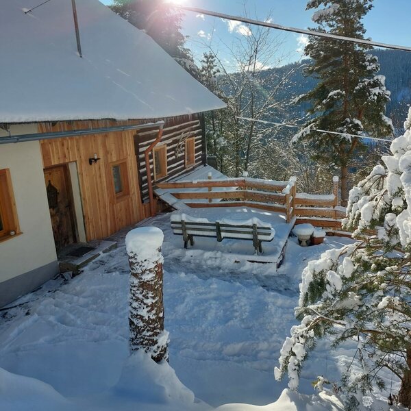 Die verschneite Alm mit Holzfassade und einer Außenterrasse mit Sitzbank, bietet einen Blick auf die winterliche Berglandschaft bei Sonnenschein.