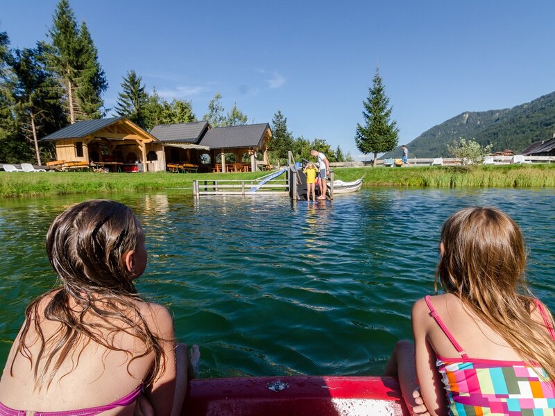 Der Badesee des Bauernhofs mit einer Badeplattform, Rutsche und Holzhütten am Ufer für Badegäste.