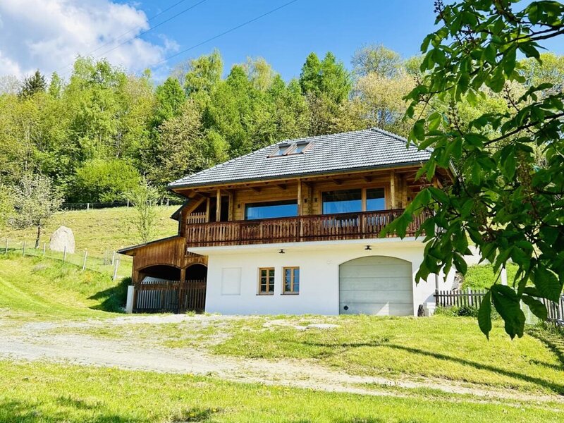 Die Außenansicht des Ferienhauses mit hölzernem Obergeschoss, Balkon und einer Garage im Erdgeschoss, umgeben von einer Wiese.