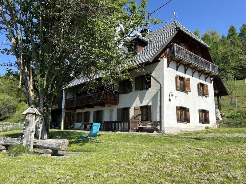Das Ferienhaus von außen mit weißer Fassade, Holzfensterläden, Balkonen, einer Rasenfläche und einem Holzbrunnen.