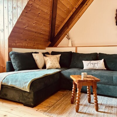 Der Wohnbereich des Ferienhauses bietet ein dunkelgrünes L-förmiges Sofa mit Kissen und Decke, einen kleinen Holztisch und ein Fenster in der holzverkleideten Dachschräge.