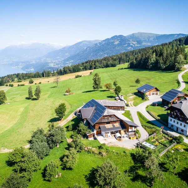 Der Bauernhof mit seinen Gebäuden inmitten grüner Wiesen, mit Blick auf einen See und die umliegende Berglandschaft.