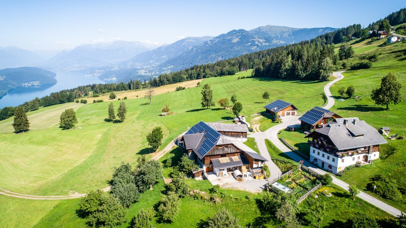 Der Bauernhof mit seinen Gebäuden inmitten grüner Wiesen, mit Blick auf einen See und die umliegende Berglandschaft.