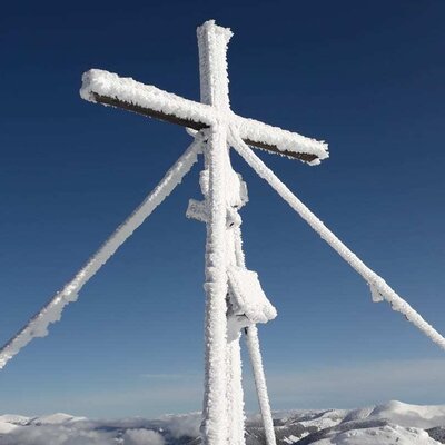 Schneebedecktes Gipfelkreuz am Mirnock unter blauem Himmel mit Blick auf die umliegenden verschneiten Berge, eine typische Winterlandschaft in der Region des Bauernhofs.