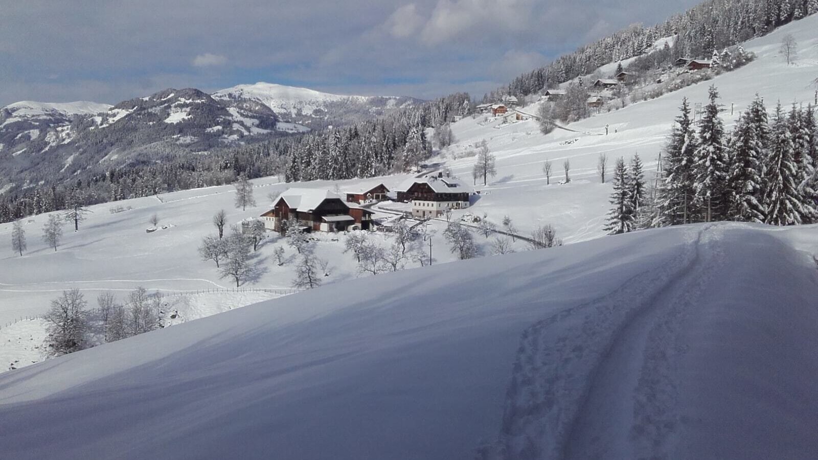 Der Bauernhof in einer verschneiten Winterlandschaft mit Bergen und Wäldern.