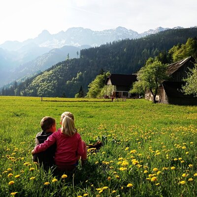 Kinder sitzen auf einer blühenden Wiese mit Blick auf den Bauernhof und die Berglandschaft.