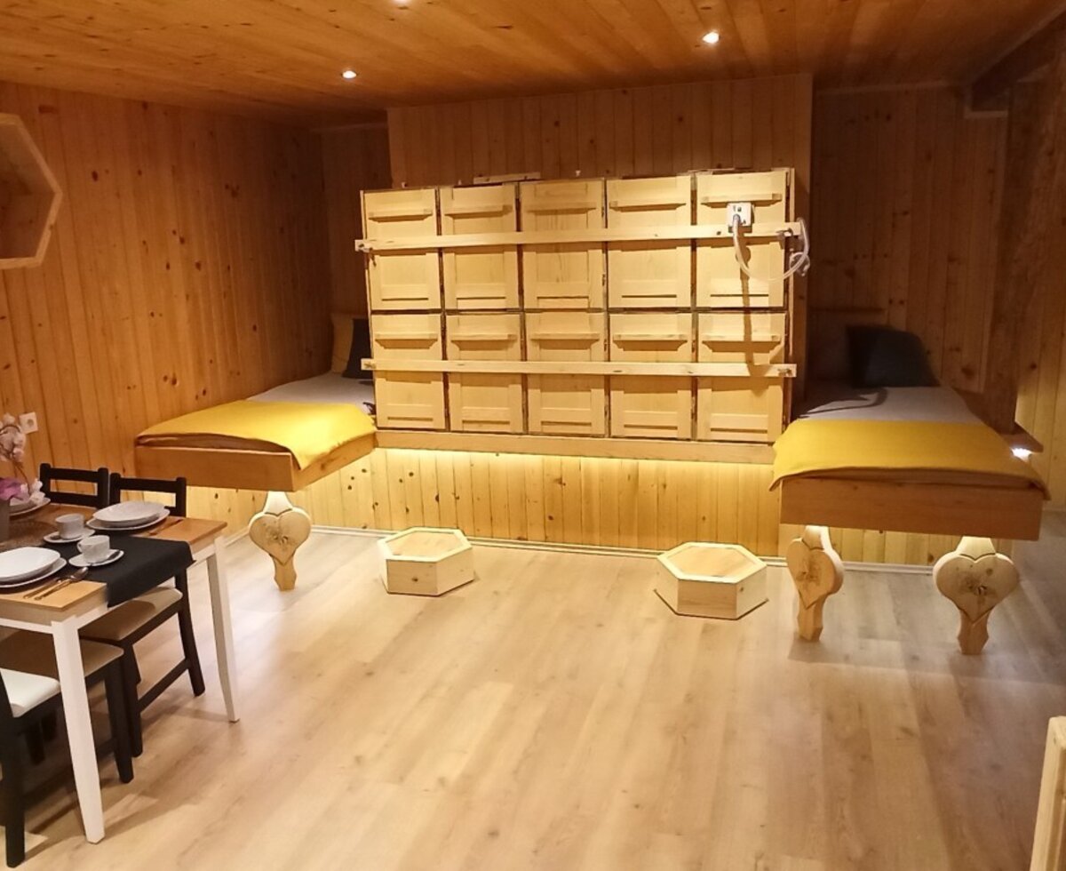 Der Raum auf dem Bauernhof zum Inhalieren von Bienenstockluft, ausgestattet mit zwei Ruheliegen, einer Wand aus Bienenbeuten und einem Esstisch.