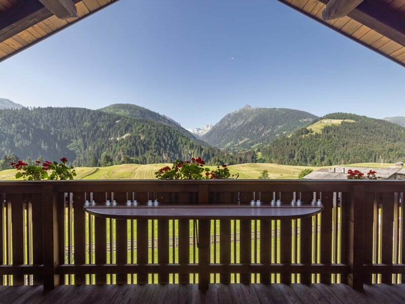 Balkon des Bauernhofs mit einem Holztisch, Blumenkästen und unverbauter Aussicht auf die umliegenden Alpen.