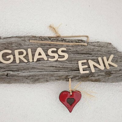 Ein Holzschild mit der Aufschrift "GRIASS ENK" und einem roten Herz heißt Gäste auf dem Bauernhof willkommen.