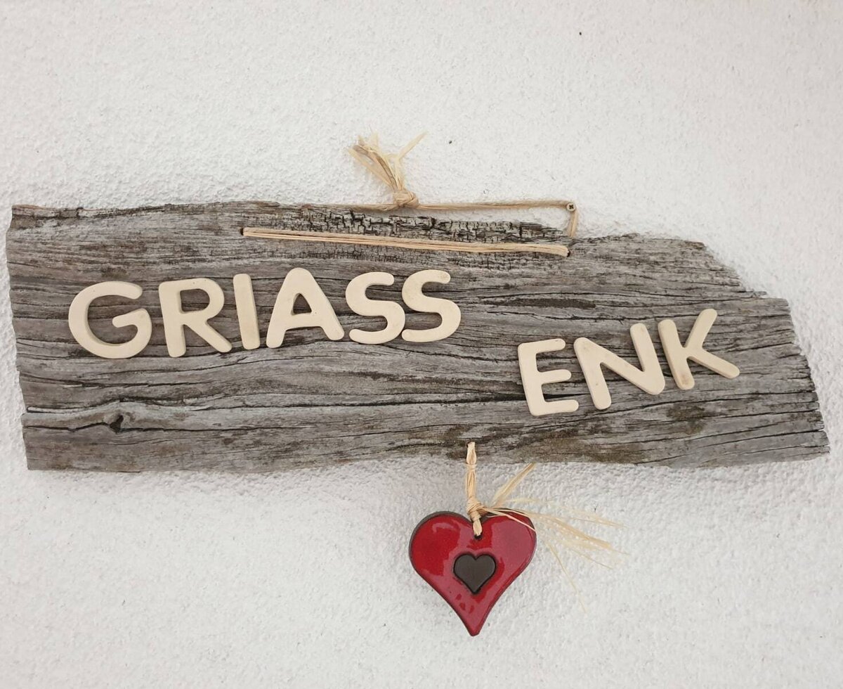 Ein Holzschild mit der Aufschrift "GRIASS ENK" und einem roten Herz heißt Gäste auf dem Bauernhof willkommen.