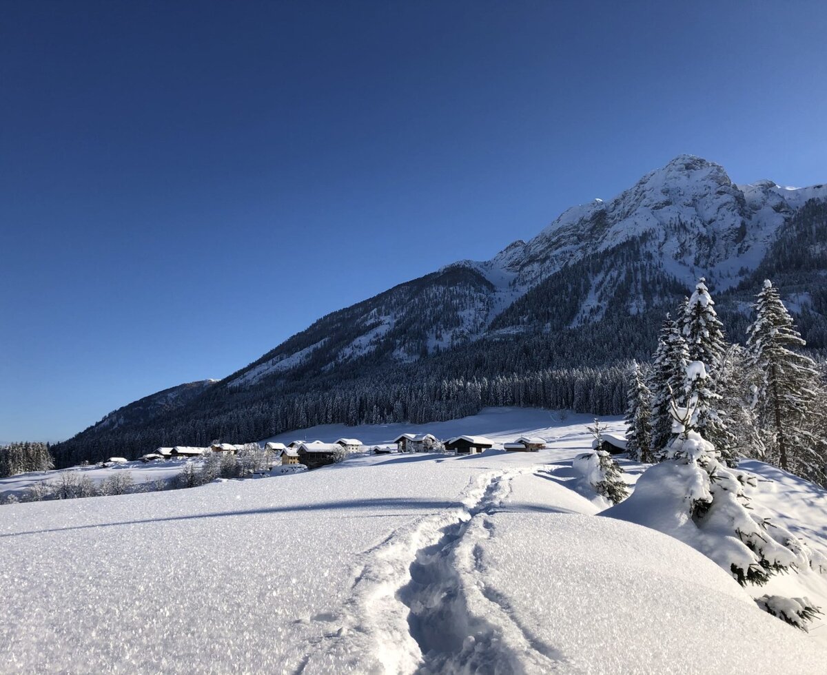 Verschneite Winterlandschaft mit Spuren im Schnee und dem Bauernhof vor den Bergen.