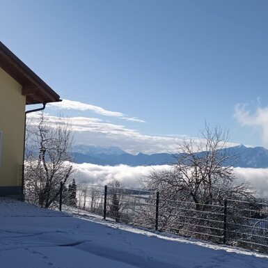 Der gelbe Bauernhof mit Garage und einer schneebedeckten Auffahrt, mit Blick auf ein Bergpanorama über einer Wolkendecke.