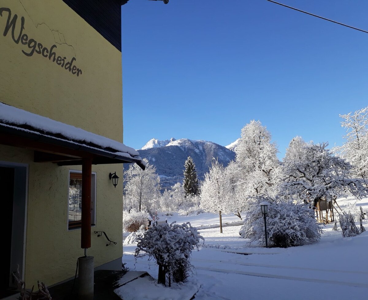 Außenansicht des Bauernhofs Wegscheider im Winter, mit verschneiter Natur und Bergkulisse unter blauem Himmel.