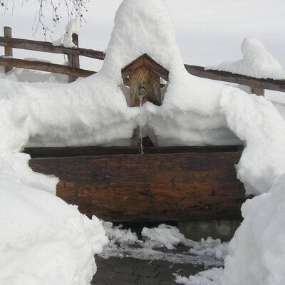 Ein verschneiter Holzbrunnen am Bauernhof mit fließendem Wasser.