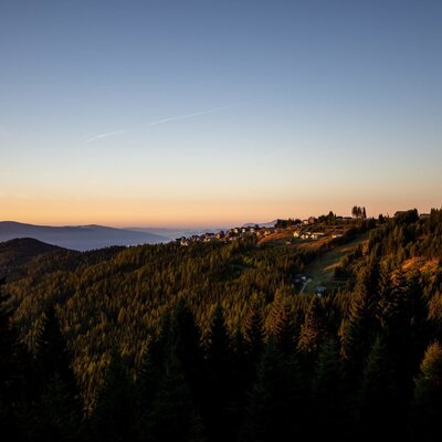 Die Alm liegt inmitten einer bewaldeten Berglandschaft mit Blick auf ein Dorf am Hang bei Sonnenuntergang.