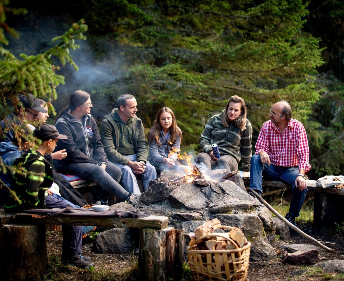 Gäste der Alm sitzen um ein Lagerfeuer im Wald, mit Holzbänken und einem Korb Brennholz.