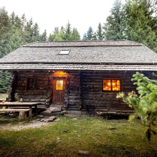 Die Alm von außen, mit Holzwänden, Schindeldach, beleuchteten Fenstern und einer Holzbank mit Tisch.