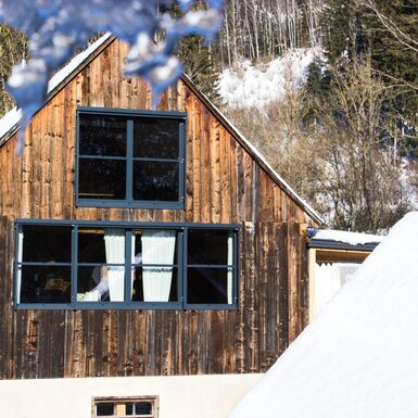 Die winterliche Holzfassade des Hofes mit großen Fenstern und einer schneebedeckten Landschaft im Hintergrund.