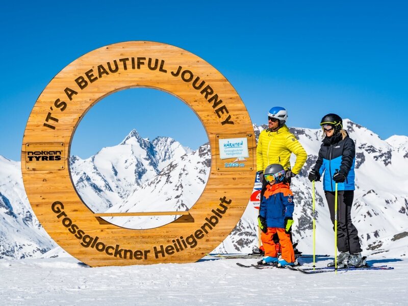 Eine Skifahrerfamilie posiert vor einem Holzring mit Bergpanorama, der die Skiregion Grossglockner Heiligenblut darstellt.