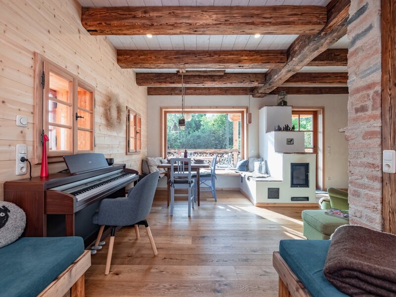Der Wohnbereich des Ferienhauses mit Klavier, Esstisch am Fenster, traditionellem Kachelofen und sichtbaren Holzbalken.