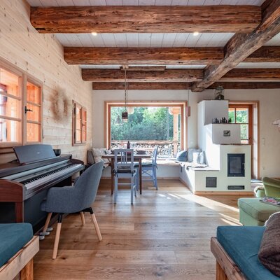 Der Wohnbereich des Ferienhauses mit Klavier, Esstisch am Fenster, traditionellem Kachelofen und sichtbaren Holzbalken.