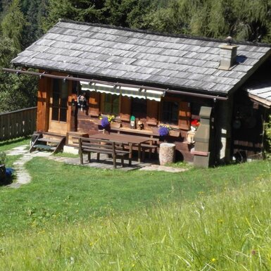 Die Alm mit Holzfassade, Steindach und einem Sitzbereich im Freien mit Holztisch und Bänken sowie einem Brunnen auf der Wiese.