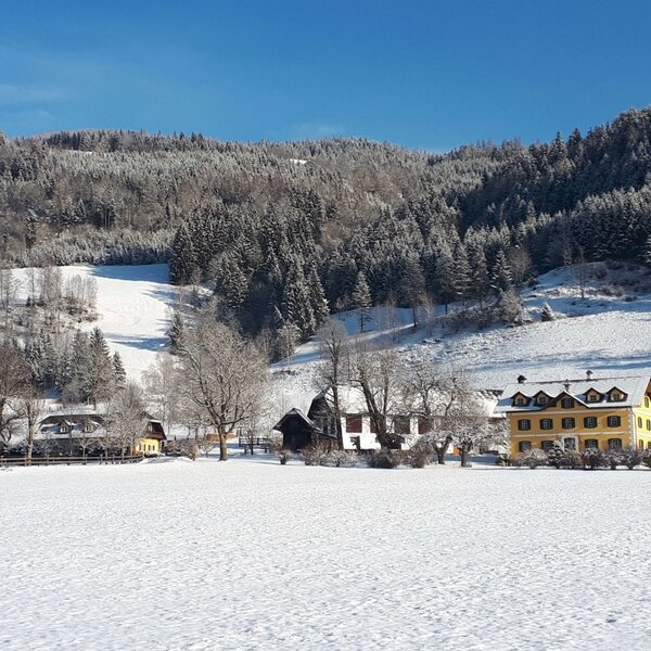 Außenansicht des Bauernhofs Fuggerhof im Winter, mit schneebedeckten Feldern und bewaldeten Hängen.