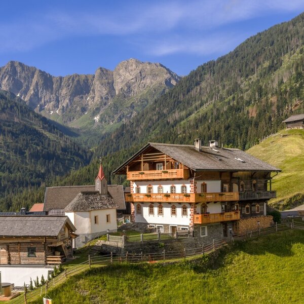 Der Bauernhof in traditioneller Holzbauweise mit mehreren Balkonen, umgeben von Bergen und grünen Hängen.