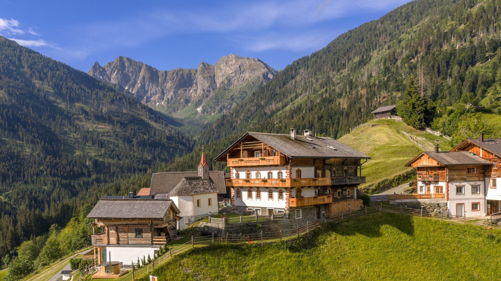 Der Bauernhof in traditioneller Holzbauweise mit mehreren Balkonen, umgeben von Bergen und grünen Hängen.