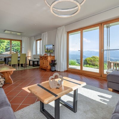 Wohnzimmer des Bauernhofs mit Kachelofen, Essbereich und großen Fenstern, die einen weiten Blick auf die Berge und den See bieten.