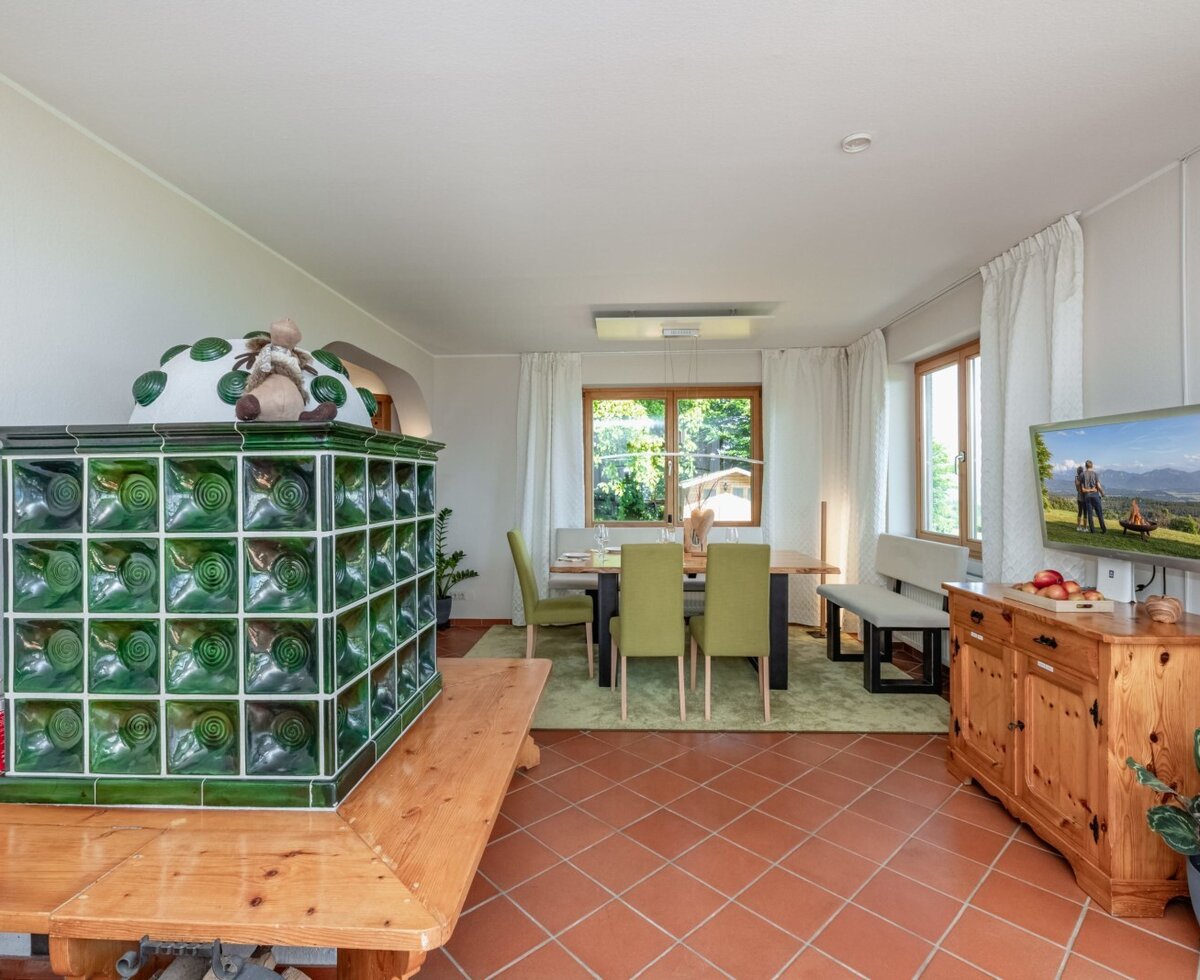 Der Wohnbereich des Bauernhofs, featuring a green tiled stove, einem Esstisch und einem an der Wand montierten TV.