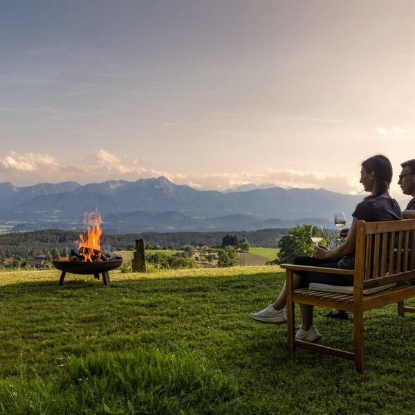 Gäste entspannen auf einer Bank am Bauernhof mit Feuerstelle und Bergblick.