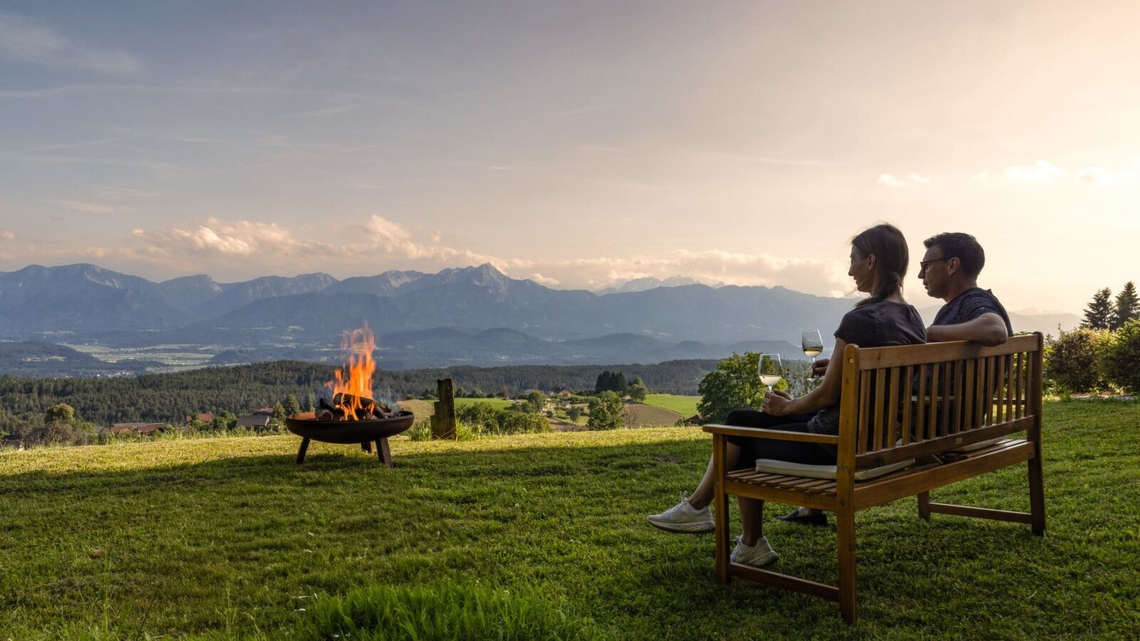 Gäste entspannen auf einer Bank am Bauernhof mit Feuerstelle und Bergblick.