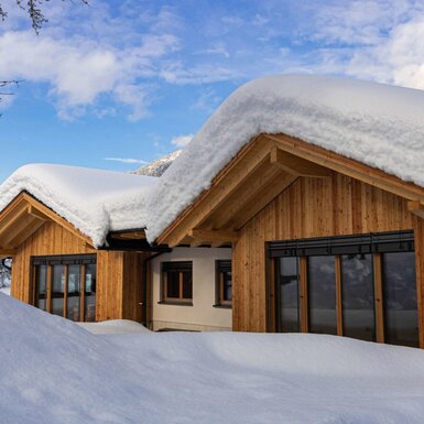 Die Außenansicht des Bauernhofs im Winter, mit Holzfassade, Fenstern mit Außenjalousien und tief verschneitem Dach.