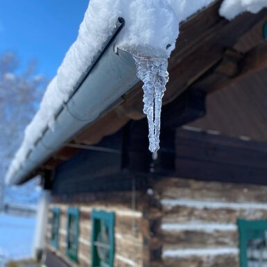 Verschneite Dachrinne der Alm mit einem Eiszapfen vor der Holzwand.