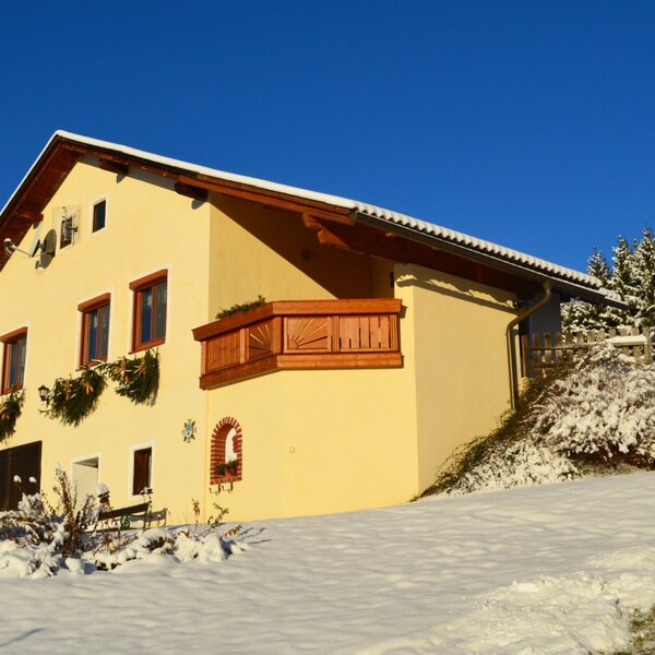Außenansicht des Bauernhofs mit gelber Fassade, Holzbalkon und verschneiter Landschaft.