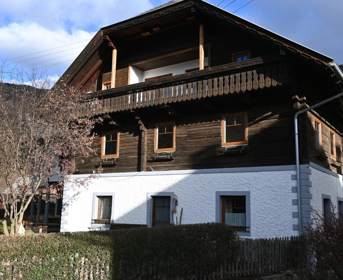Außenansicht der Ferienwohnung mit weiß verputztem Erdgeschoss, Holzbalkon und Holzfassade im Obergeschoss.