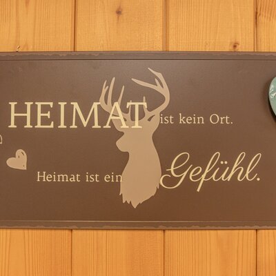 Dekoratives Holzschild mit dem Spruch "Heimat ist kein Ort. Heimat ist ein Gefühl.", Hirschmotiv und einem dekorativen Herz am Bauernhof.