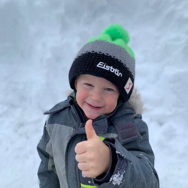 Ein Kind in Winterkleidung gibt im Schnee auf dem Bauernhof einen Daumen hoch.