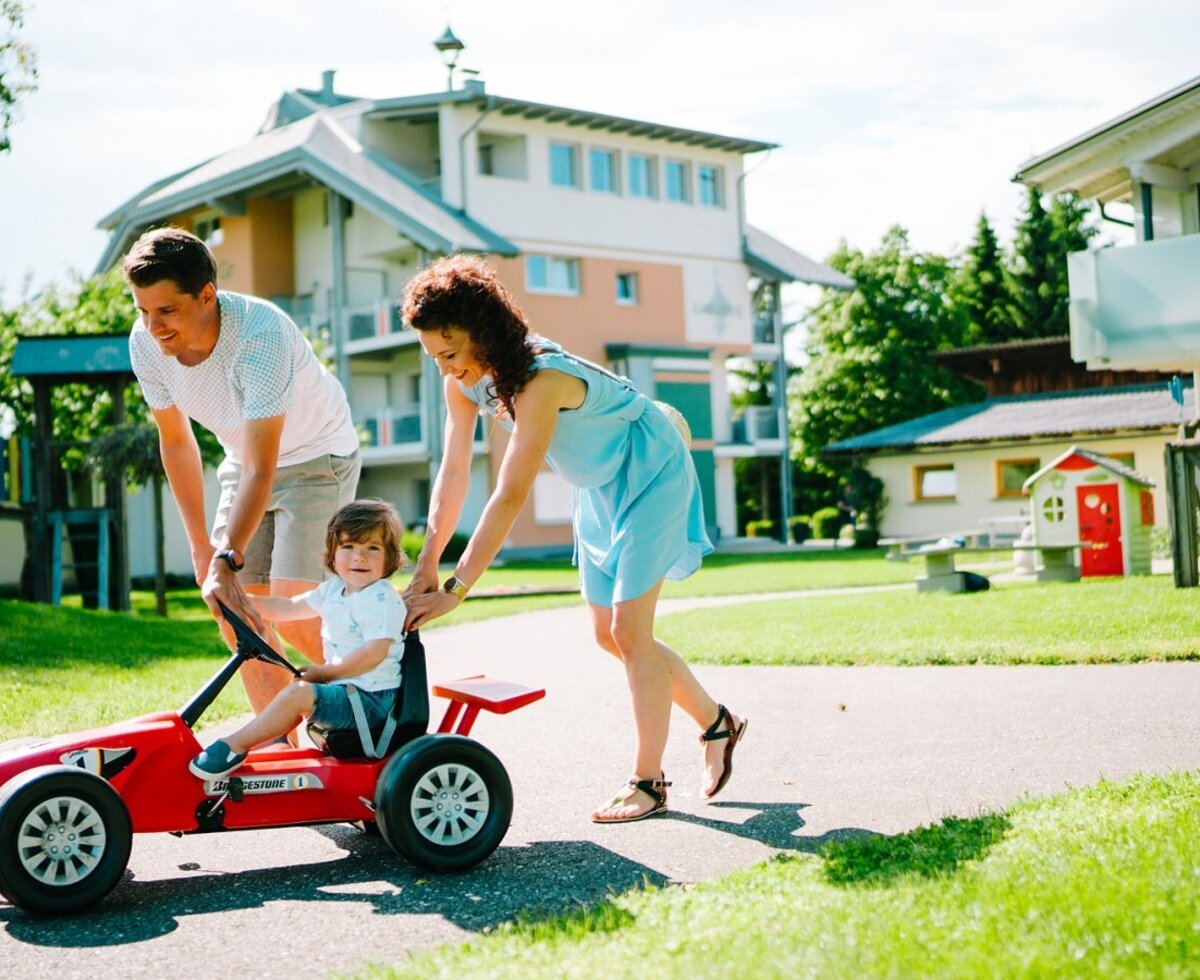 Familie spielt mit einem Tretfahrzeug auf dem Gelände des Bauernhofs, mit Spielplatz im Hintergrund.