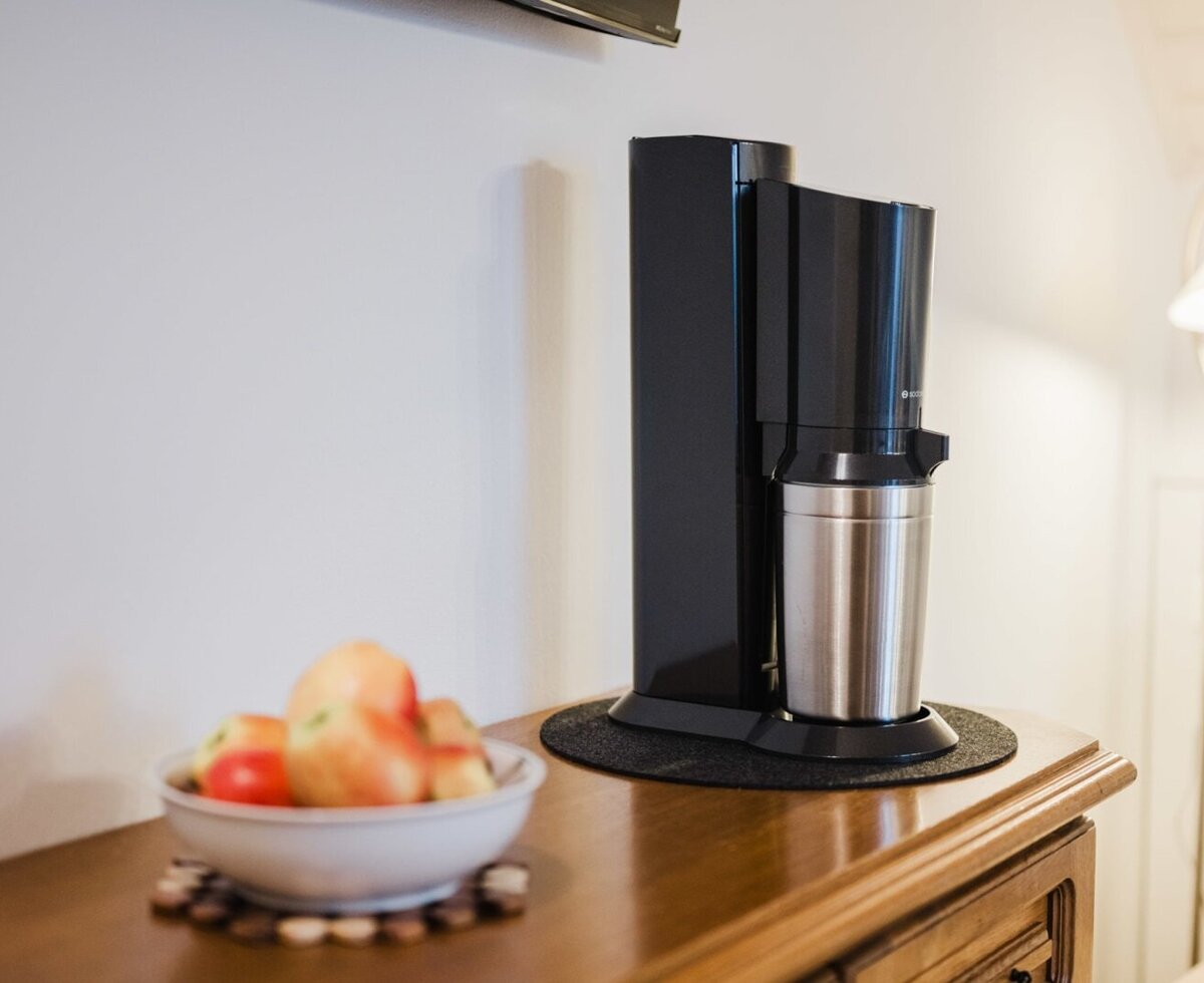 SodaStream-Gerät mit Glasflaschen, daneben eine Schale mit Äpfeln.