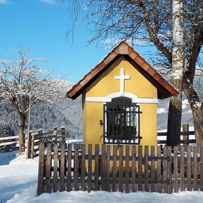 Der besondere Kraftort "das geweihte Brünnl" als gelber Schrein mit Kreuz in der verschneiten Berglandschaft.