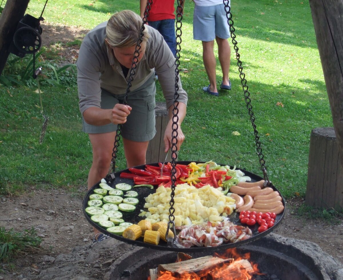 Kochen am Lagerfeuer mit einer großen Pfanne voller frischer Gemüse, Würstchen und Kartoffeln auf dem Bauernhof.