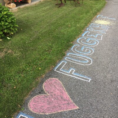 Der Weg zum Bauernhof mit Kreidezeichnungen, die ein Herz und den Schriftzug "Fuggerhof" zeigen, neben einem Rasen mit Blumen und einem historischen Bauernhofgerät.