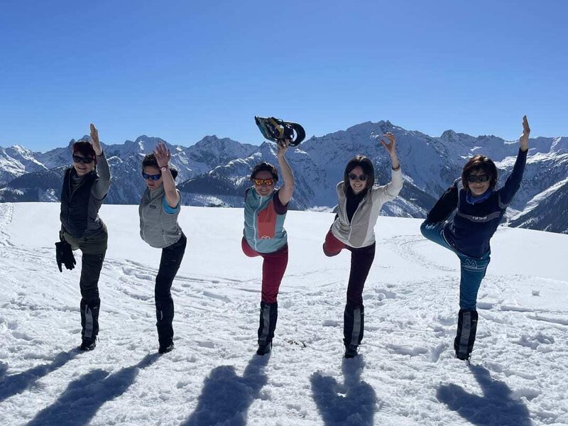 Fünf Personen in Yoga-Posen im Schnee vor einer Bergkulisse.