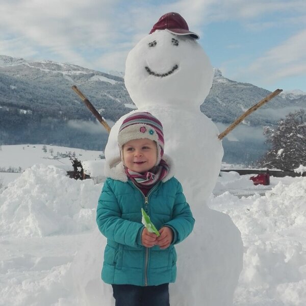 Ein Kind steht neben einem Schneemann im Schnee auf dem Bauernhof, mit Bergen im Hintergrund.