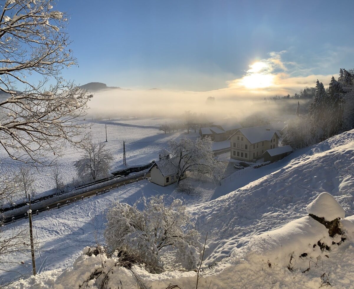 Der verschneite Bauernhof Gschlosserhof in einer idyllischen Winterlandschaft mit sanftem Sonnenschein und Nebelschwaden.